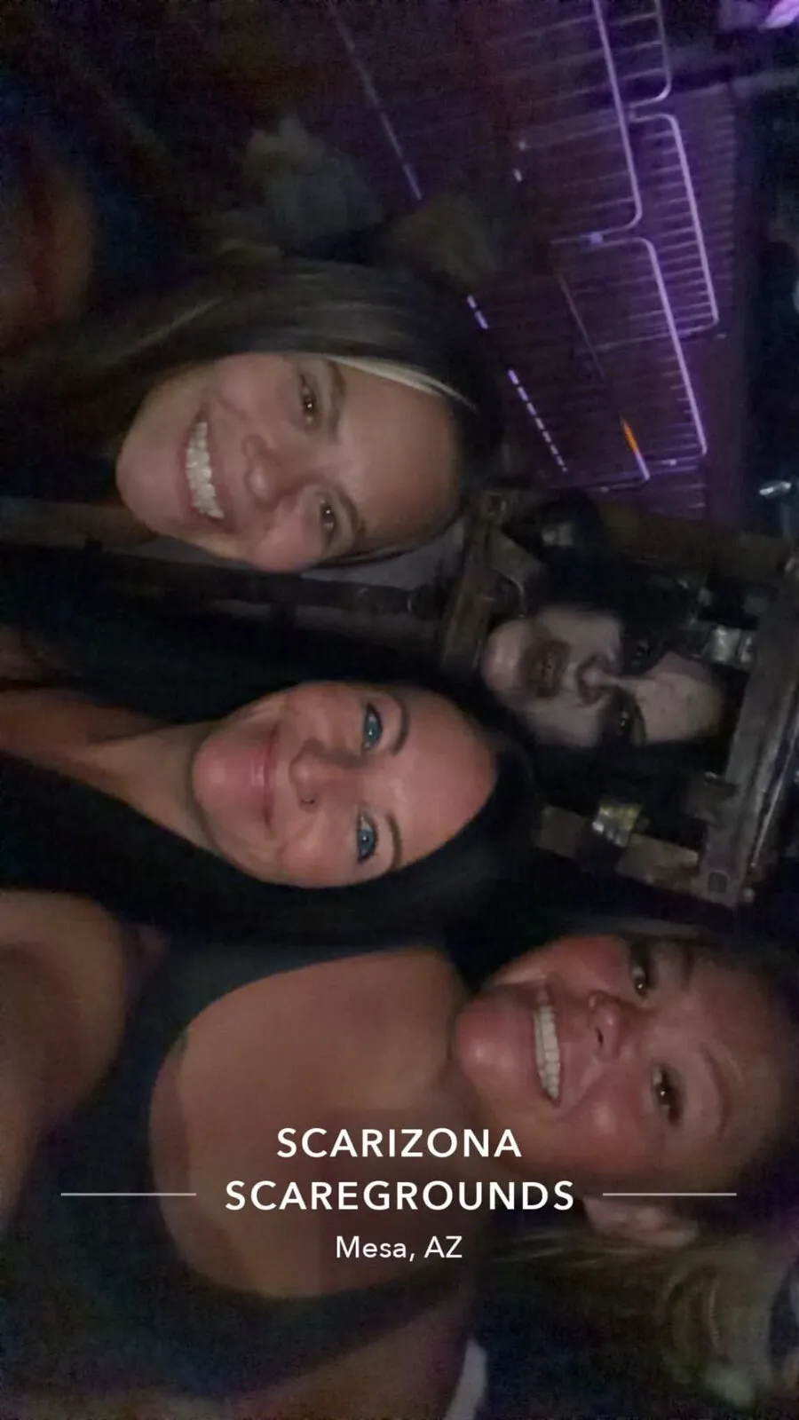 Three smiling guests take a selfie while a masked character looms behind them at Scarizona Scaregrounds