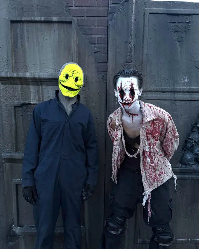 A yellow smiling-mask scare actor beside a bloodied clown character against steel doors at Scarizona Scaregrounds