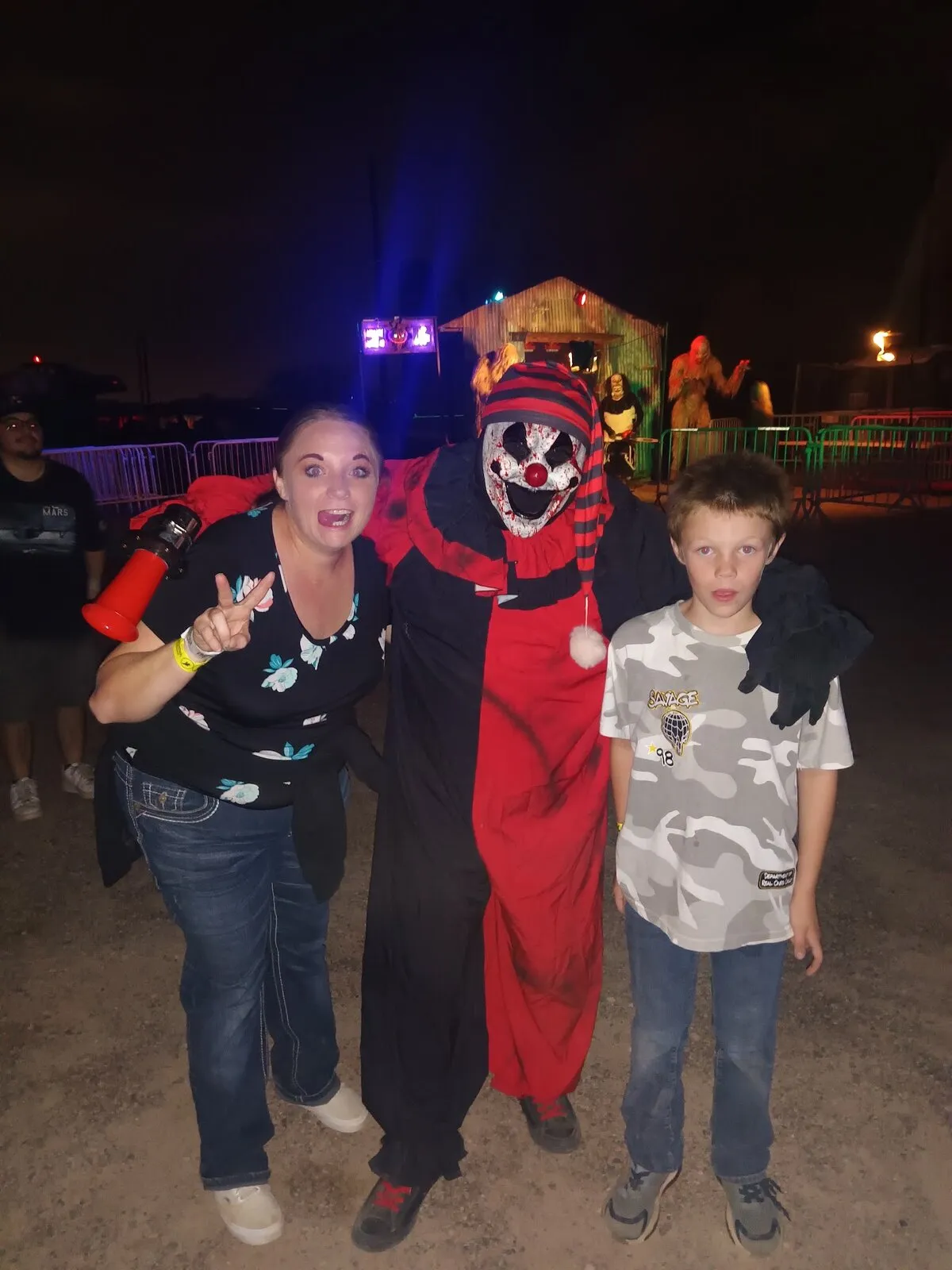 A guest and a young boy pose with a grinning jester clown in red and black on the Scarizona midway